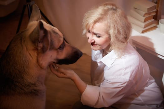 Adult mature woman of 40-60 with big shepherd dog in white shirt. Room with calm cozy evening atmosphere with transparent curtains and soft warm light of lamps. Concept of love for animals and pet