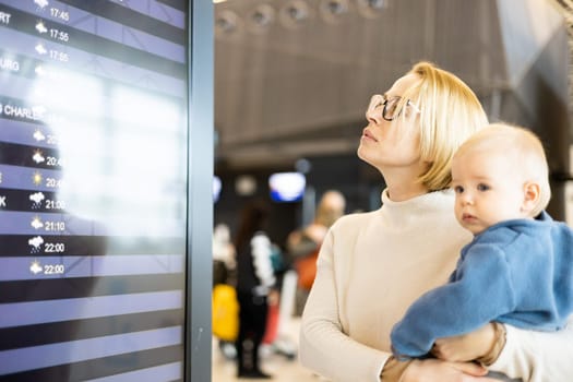 Mother traveling with child, holding his infant baby boy at airport terminal, checking flight schedule, waiting to board a plane. Travel with kids concept