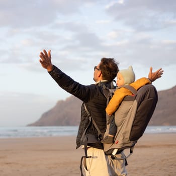 Father rising hands to the sky while enjoying pure nature carrying his infant baby boy son in backpack on windy sandy beach of Famara, Lanzarote island, Spain at sunset. Family travel concept