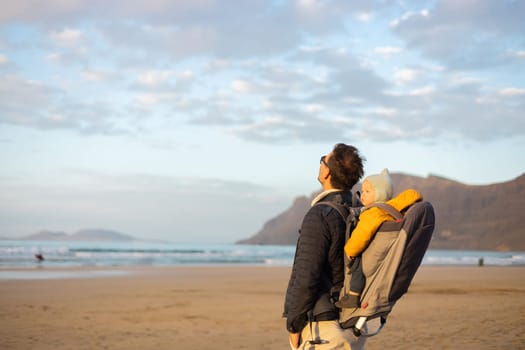 Father rising hands to the sky while enjoying pure nature carrying his infant baby boy son in backpack on windy sandy beach of Famara, Lanzarote island, Spain at sunset. Family travel concept