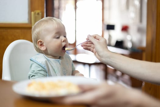 Mother spoon feeding her infant baby boy child sitting in high chair at the dining table in kitchen at home.