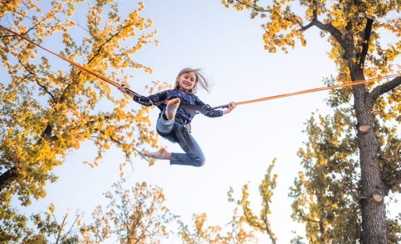 Bottom view of little girl on trampoline jumping ropes on blue sky and autumn treetops background