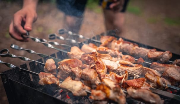 Man hand straightens the skewer with barbecue delicious grilled meat on brazier outside at summertime. Person preparing pork steaks with smoke during picnic at the nature. Closeup view at hot BBQ