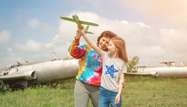 Mother and daughter launching toy plane at the field with real airplane on background. Girl and woman spending family time together at the nature