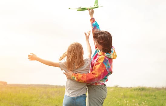 Mother and daughter launching toy plane at the field. Girl and woman spending family time together at the nature. Portrait from back with sunset rays