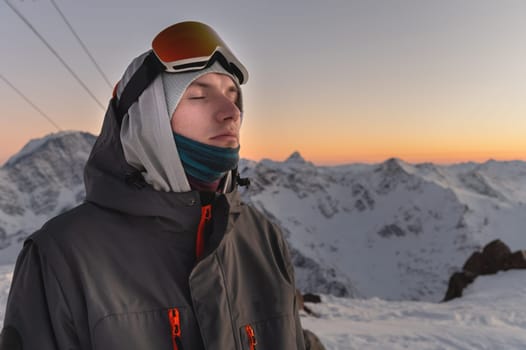 a man in ski goggles against the backdrop of snow-capped mountains and blue sky during sunset. eyes closed, enjoyment and happiness from silence and outdoor recreation.