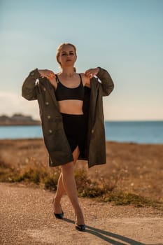 Portrait blonde sea cape. A calm young blonde in an unbuttoned khaki raincoat walks along the seashore, under a raincoat a black skirt and top.