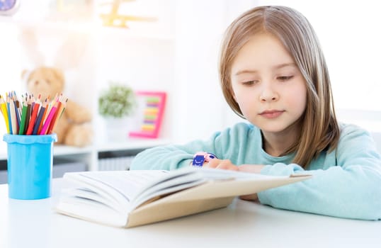 Cute little girl reading book on desk in bright room