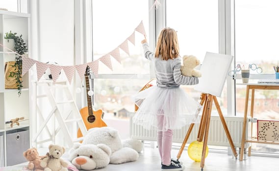 Cute little girl pointing at window for teddy bear in playroom