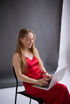 A happy business woman blogger smile in a red dress is working, typing on a laptop apple and sitting on a chair. portrait blonde assistant of hands with computer MacBook . Vertical