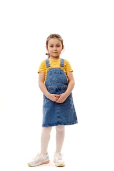Full length vertical studio portrait of a Caucasian beautiful little child girl wearing blue denim sundress and yellow t-shirt, looking at camera, standing isolated on white background with copy space