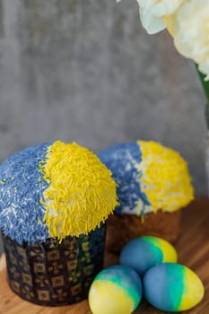 Delicious Easter cakes in the colors of the flag of Ukraine, yellow-blue colored Easter eggs on a wooden table with flowers in the background. place for text. selective focus