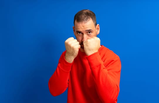 Bearded Hispanic man in his 40s wearing a red jersey standing guard ready to start a fight with his fists, isolated over blue background