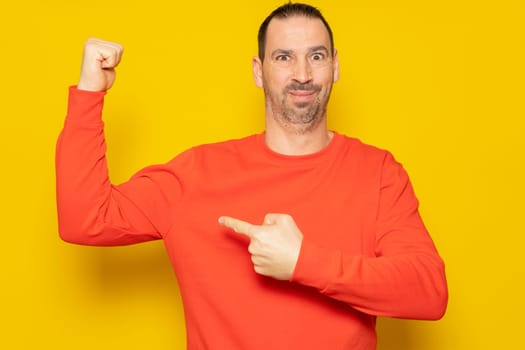 Portrait bearded latino man pointing finger at arm muscle, discount, looking at camera with satisfied face. Indoor studio shot isolated on yellow background