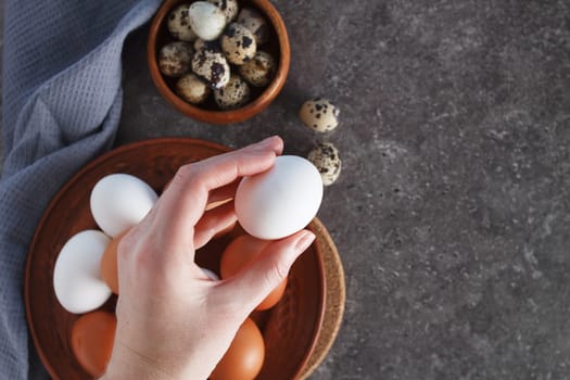 A woman's hand holds a chicken egg over a plate of eggs on the kitchen table.
