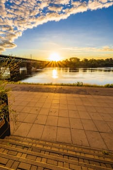 A beautiful sunrise over a wide river next to a long green bridge seen from the steps of the city promenade