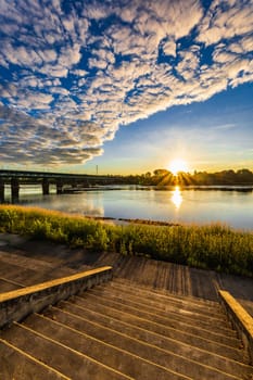 A beautiful sunrise over a wide river next to a long green bridge seen from the steps of the city promenade