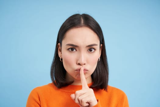 Close up of angry brunette asian girl, shush, touches lips with finger, tells to be quiet, stands over blue background.