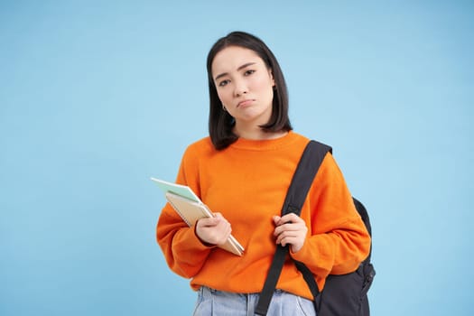 Sad sulking asian student, girl with backpack and notebooks, looking upset, standing over blue background. Copy space