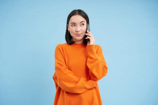 Korean girl in orange sweater talks on mobile phone, speaks on cellphone with happy smiling face, orders delivery, consults with someone on telephone.