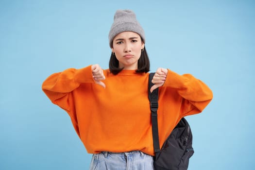 Upset asian woman shows thumbs down, dislike something, looking frustrated and disappointed, standing over blue studio background.