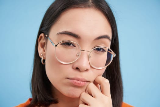 Close up portrait of asian woman looks intrigued, wears glasses, squints thoughtful, thinking, making assumption, standing over blue background.