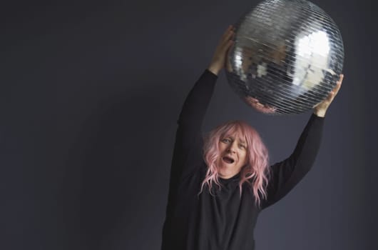 A man in a black turtleneck in a pink wig is dancing holding a mirrored disco ball in his hand. Close-up.