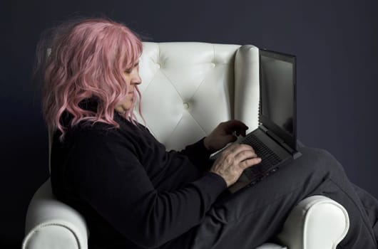 A man in a pink wig sits in a white chair and works at a computer.