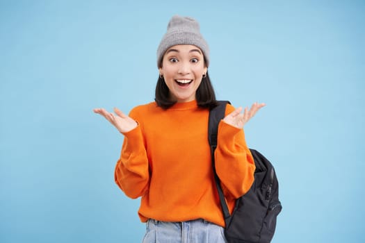 Happy asian woman in warm hat, claps hands from surprise, laughs and looks amazed, excited by smth, stands over blue background.