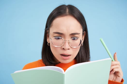 Woman in glasses looking in her notebook with serious face, making notes, writing down information with thoughtful expression, blue background.