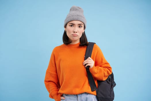 Young asian woman in hat, holds backpack, sulks and looks confused, perplexed emotion, stands over blue background.
