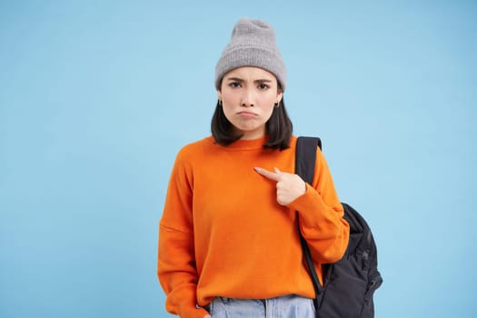 Sad asian girl points at herself with upset, gloomy face expression, stands over blue studio background.