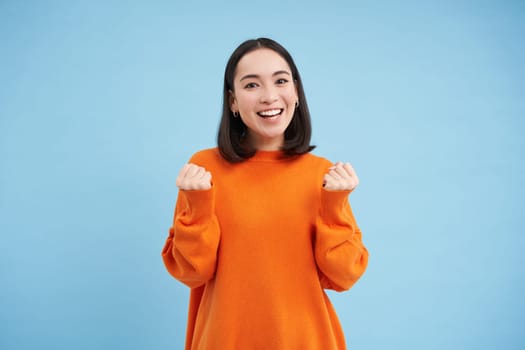 Excited young woman celebrating, jumping with hooray smiling face, winning, achieve goal, triumphing while standing over blue background.