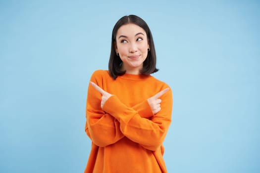 Decisions. Young asian woman points sideways, shows left and right with indecisive smiling face, choosing between two options, stands over blue background.