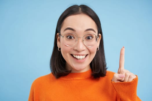 Portrait of smiling brunette asian woman in glasses, raises one finger, eureka sign, pitching an idea, has revelation, stands over blue background.