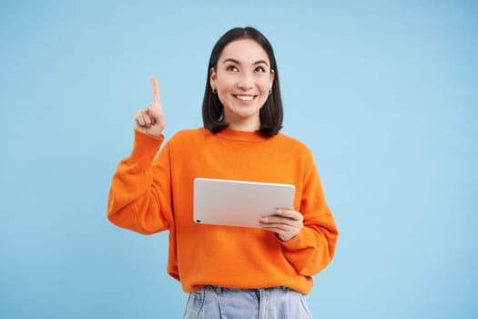Beautiful young korean woman with tablet, pointing finger up and looking on top advertisement, showing banner, standing over blue background. Online shopping concept.