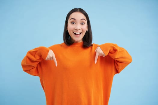 Cheerful asian woman shows advertisement below, points down with smiling face, inviting people to click on link, follow arrow, standing over blue background.