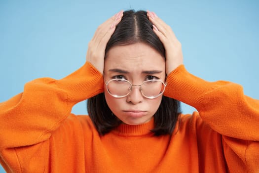 Portrait of asian woman in glasses, holds hands on head complicated face, feels troubled, has problems, standing confused over blue background.