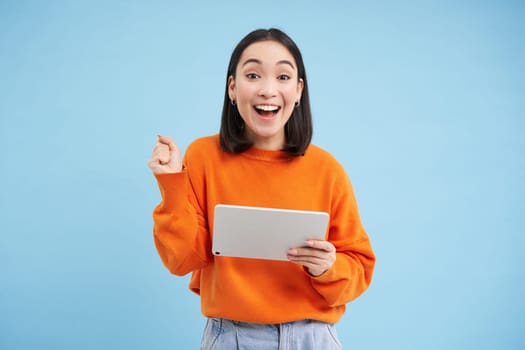 Amazed korean woman with digital tablet, looks surprised and excited, standing over blue background.