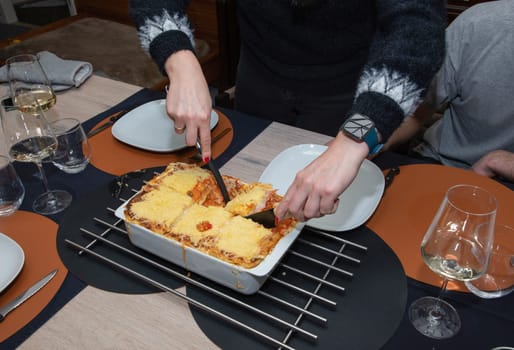 a woman serving hot homemade lasagna on plates to a family member sitting at the table, High quality photo