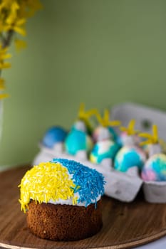Easter composition with yellow-blue Easter cakes, sprinkles, Easter multi-colored eggs. wooden stand and spring flowers on a yellow background. copy space.