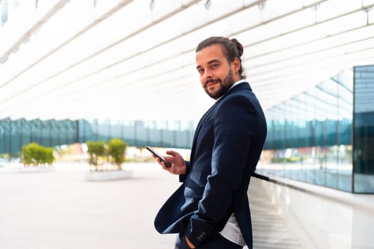 Businessman standing outdoor use smartphone looking camera screen confident. Handsome hispanic male business person in business suit holding smartphone in hand. Modern business person low angle view