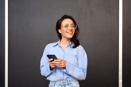 Portrait smiling woman holding smartphone in hand looking away, wear glasses, types text message on mobile phone, enjoys online communication, on black studio background. Communication concept