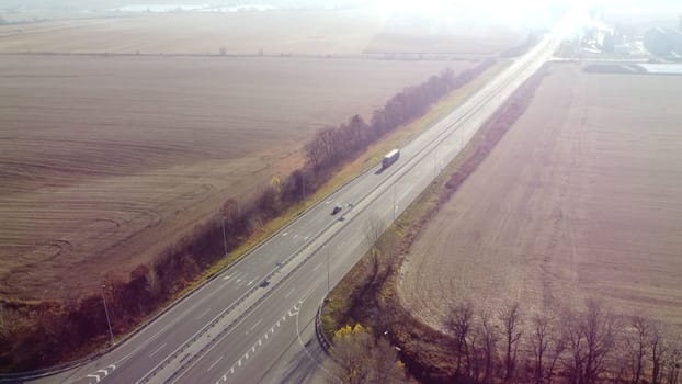 Beautiful top view of automobile road with different cars driving along it between agricultural fields on a sunny autumn day. Highway, roadway in nature. Sun glare light, highlight. Aerial drone view