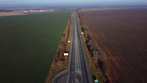 Beautiful panoramic landscape. Black paved highway with white markings,many driving passing cars, large agricultural fields with green agricultural crops and plowed black soil on a sunny autumn day.