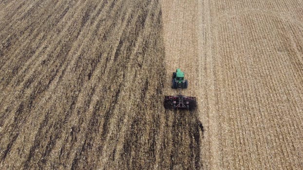 Tractor plowing the ground. Flying over green tractor that plows up ground in yellow field after harvesting wheat on autumn day. Tractor digging land. Agricultural work on field. Aerial drone view