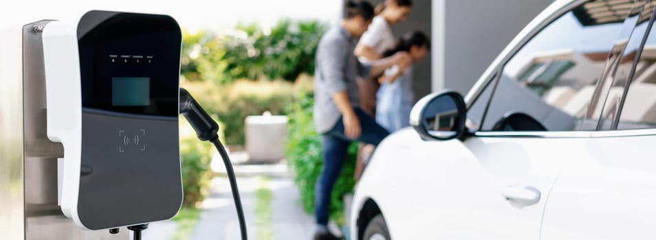 Focus closeup electric vehicle recharging battery from home electric charging station with blurred family in background. Renewable clean energy car for progressive eco awareness lifestyle concept.