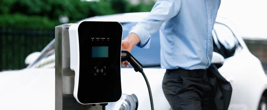 Closeup progressive suit-clad businessman with his electric vehicle recharge his car on public charging station in modern city with power cable plug and renewable energy-powered electric vehicle.