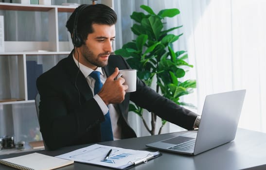 Manager of call center operator office sitting on his desk with his coffee while working on laptop. Office worker wearing headset and black suit working on customer support or telemarketing. fervent