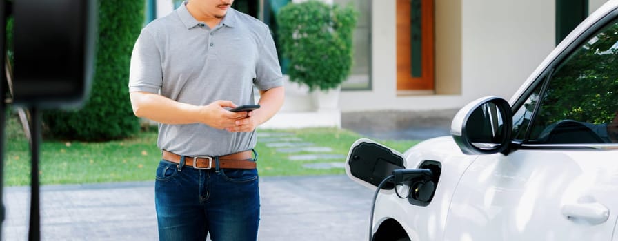 Progressive asian man install cable plug to his electric car with home charging station in the backyard. Concept use of electric vehicles in a progressive lifestyle contributes to clean environment.